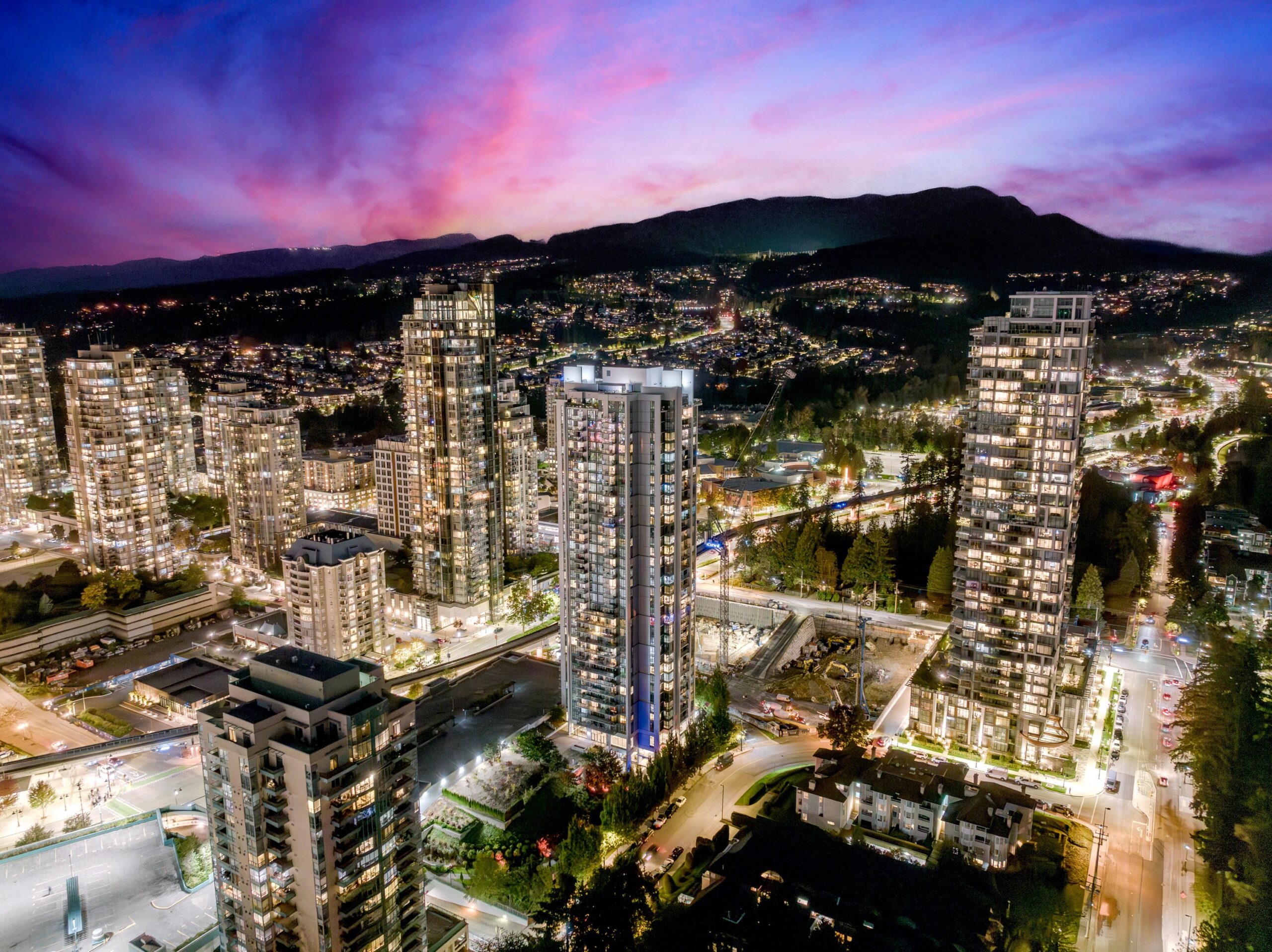 Elevated night drone photography of the Coquitlam City Centre skyline at sunset, showcasing high-rise towers, urban density, and mountain silhouettes.